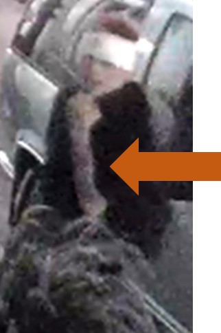 Person of interest wearing a dark top, white cloth wrapped are their head, carrying a bag, leaning against a vehicle.